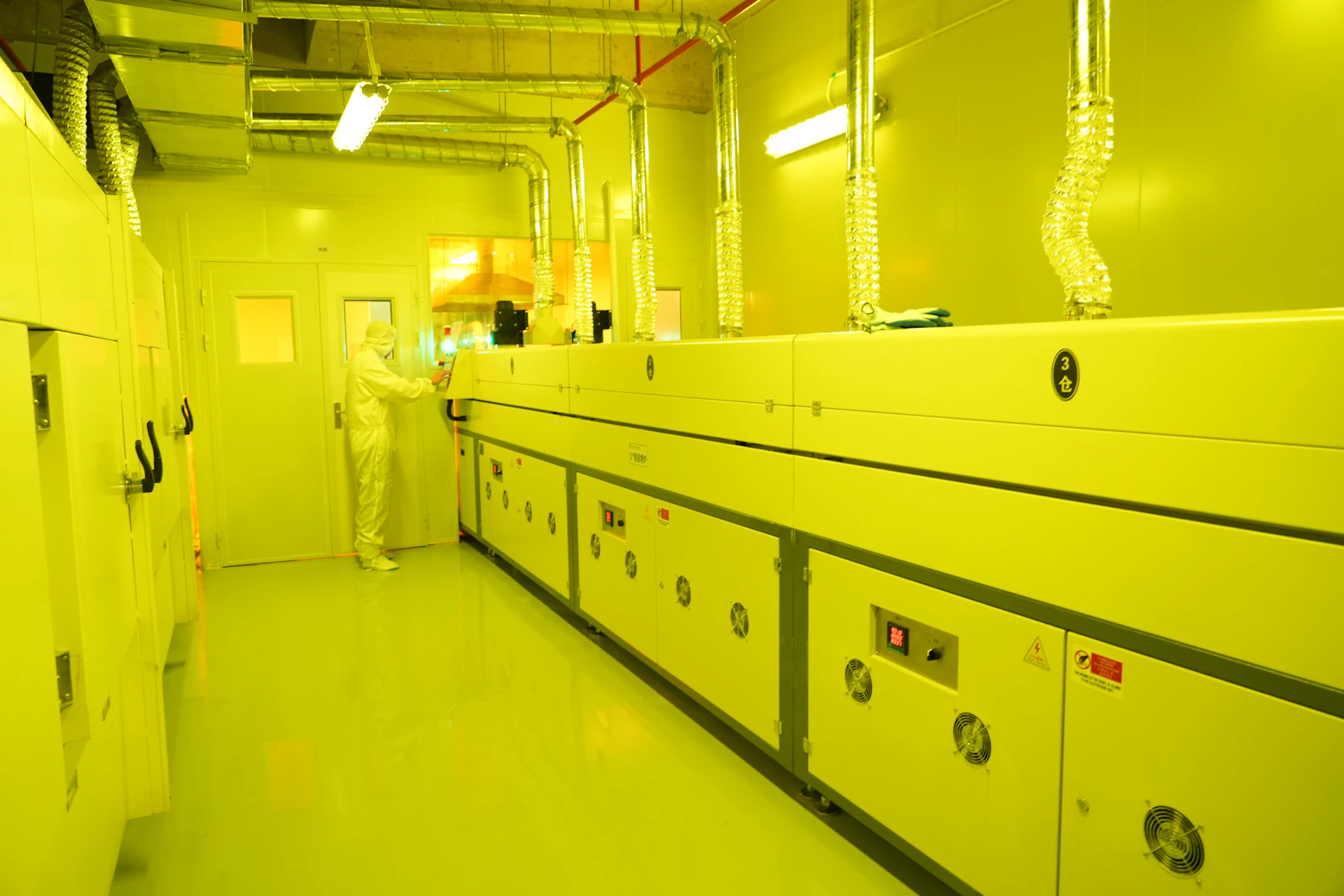 Technician in a cleanroom suit operating semiconductor equipment under yellow light